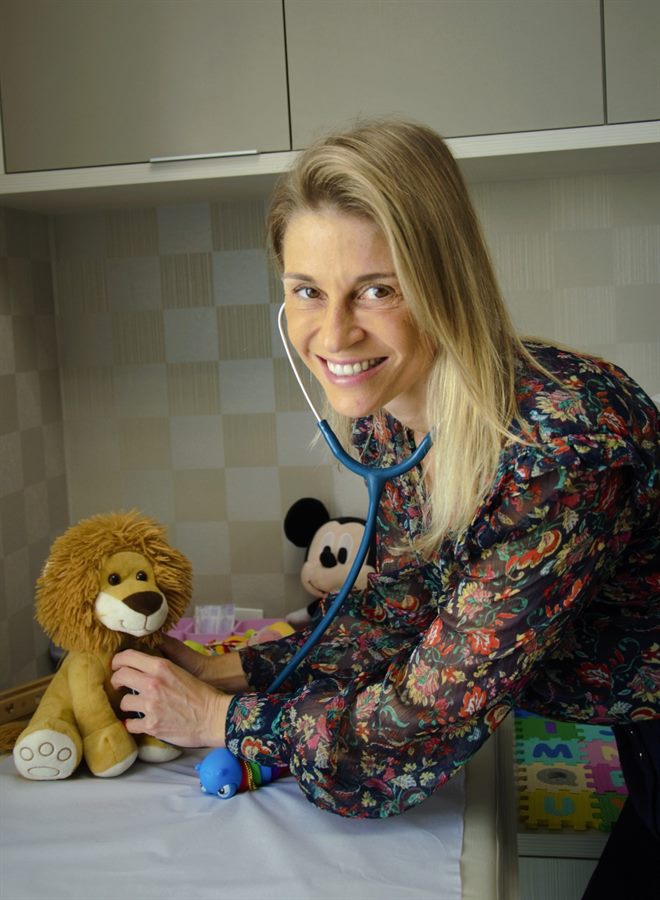 Dra. Milena Catani sorrindo no consultório, ao lado de um leão de pelúcia
